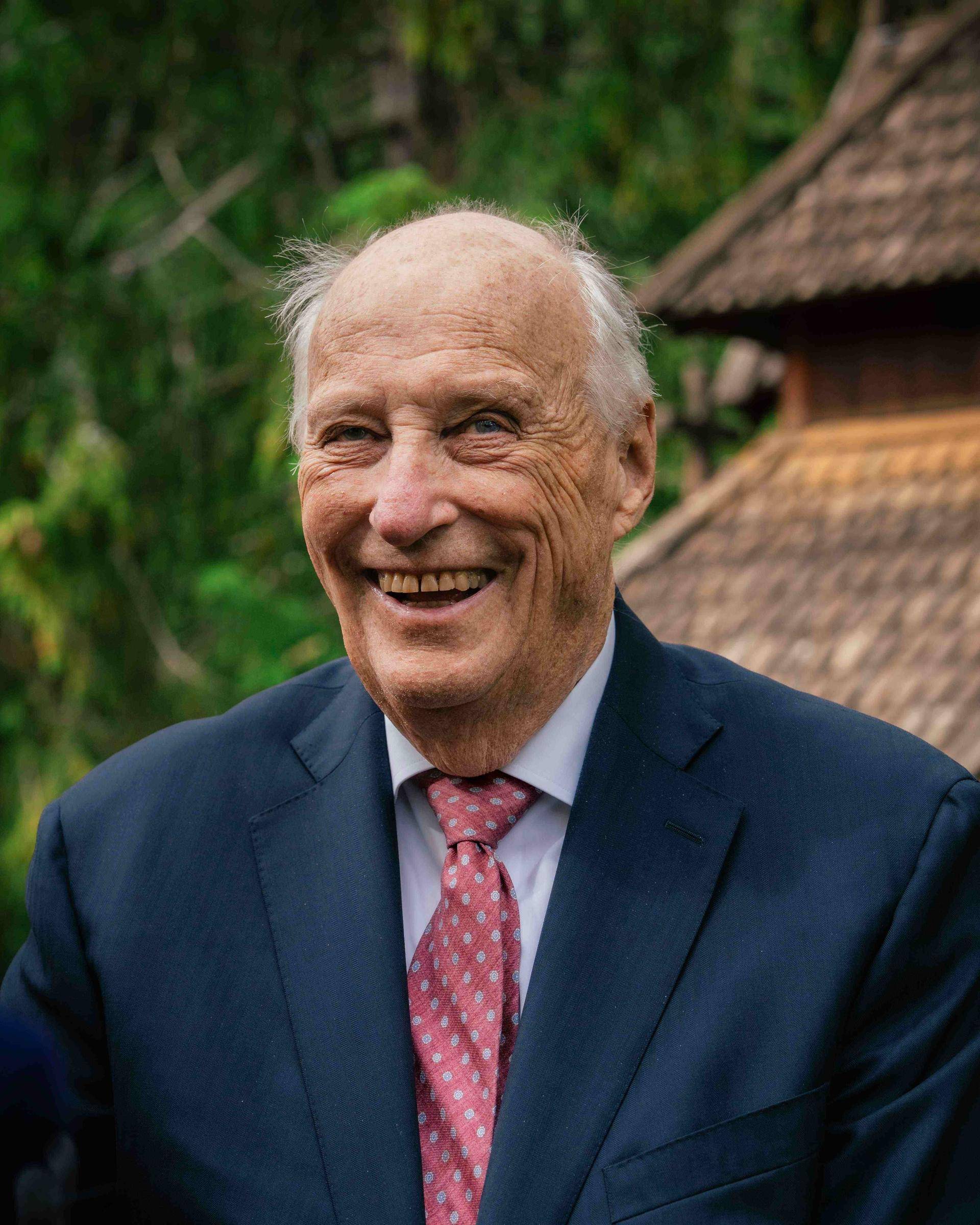 Eldre mann smiler bredt mot kamera, med grønn skog og en trebygning i bakgrunnen. Kong Harald V.